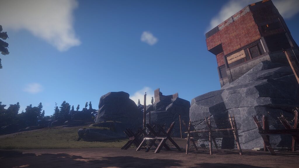 A screenshot from the game Rust, where you can see a base built on top of a cliff, and the base being built of scrap metal. Below the cliff there's barb wire fences.